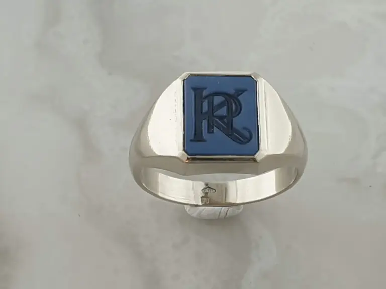Close-up of a silver ring with a blue square top featuring the letter 'R' and an ornate monogram design.