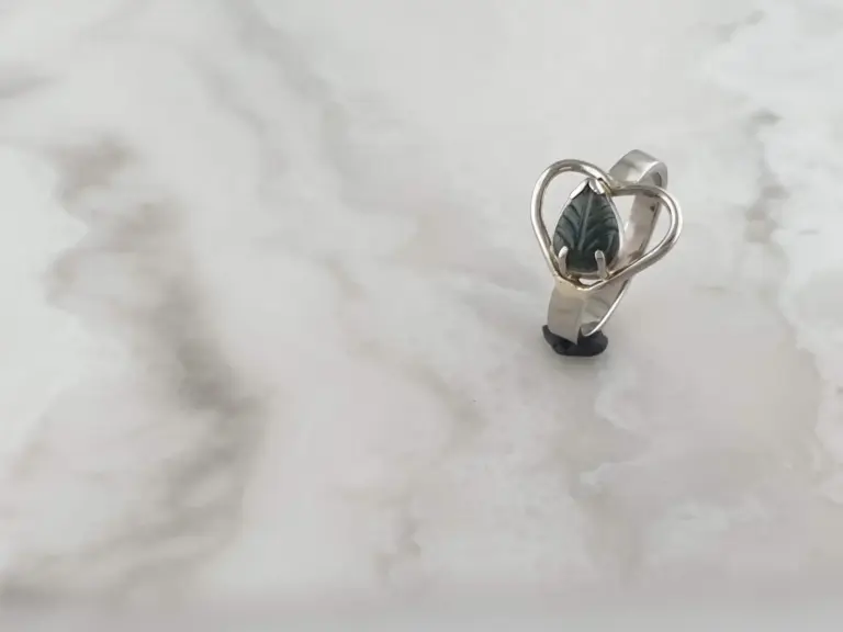 A silver ring with a heart-shaped design encasing a dark gemstone, placed on a light-colored, textured surface.