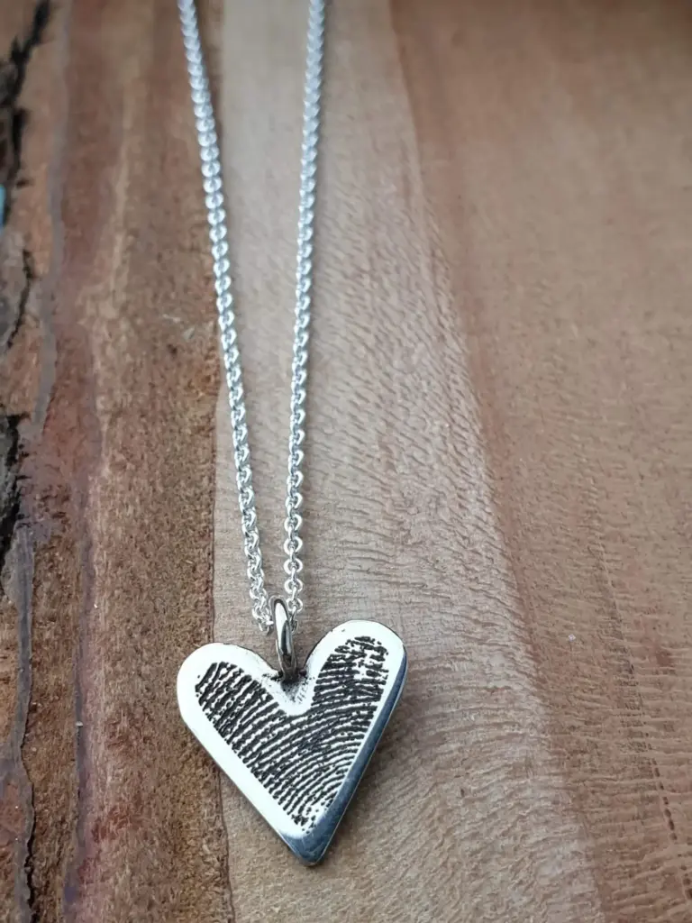 A silver necklace with a heart-shaped pendant engraved with fingerprint patterns, resting on a wooden surface.