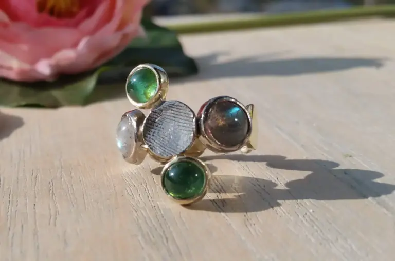 Close-up of a stack of colorful rings with various gemstones, placed on a wooden surface with a large pink flower and green leaves in the background.