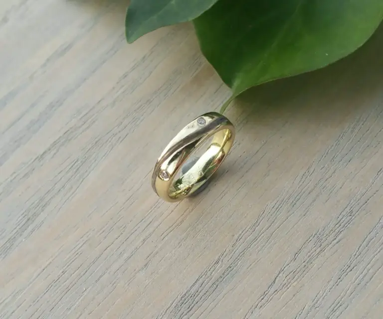 A gold ring with embedded diamonds rests on a light-colored wooden surface near a large green leaf.