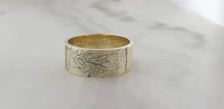 A wide silver ring with a textured engraved pattern on its surface, resting on a white surface.