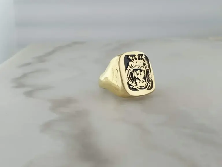 A gold ring with an engraved crest or emblem on top, resembling a shield with intricate designs, placed on a white surface.