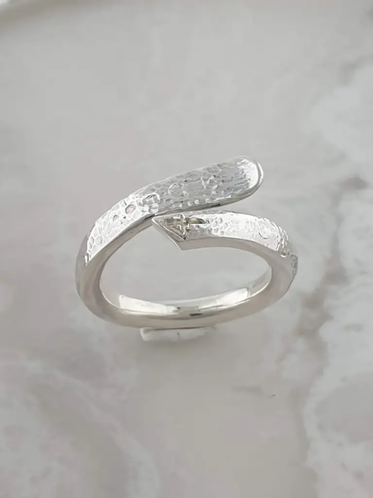 Close-up of a silver ring with an open design, textured surface, and engraved details, resting on a plain white background.
