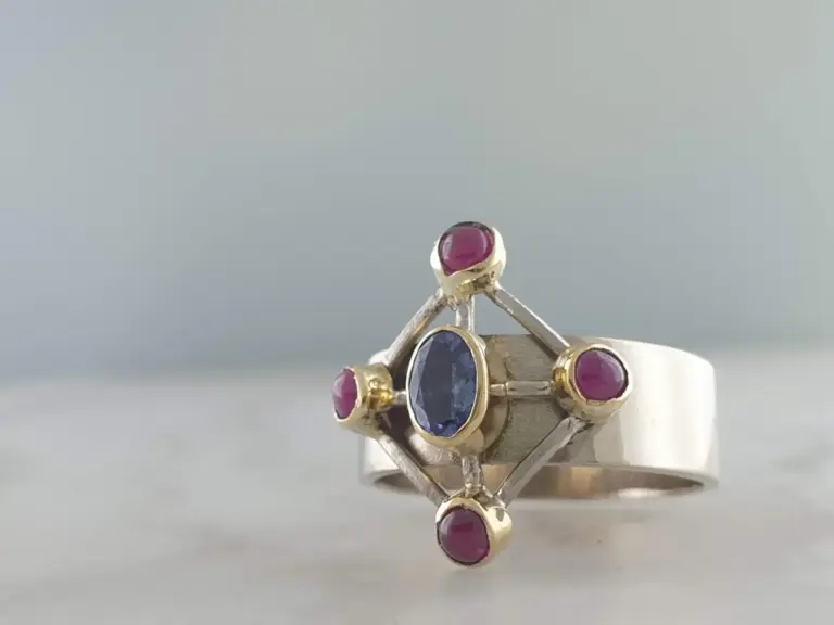 Close-up of a silver ring with multiple gemstones, including a large blue gemstone and several smaller pink gemstones, set in a geometric design.