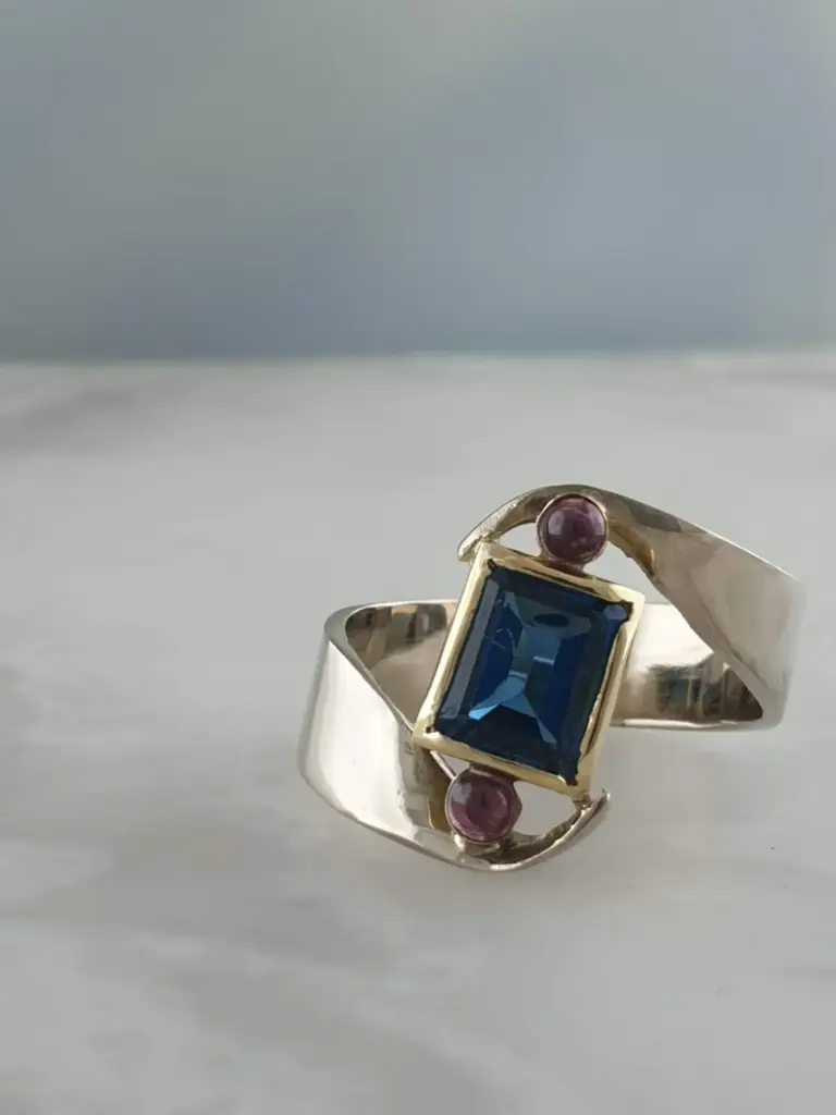 Silver ring with a large square blue gemstone surrounded by two small purple stones, placed on a white surface.