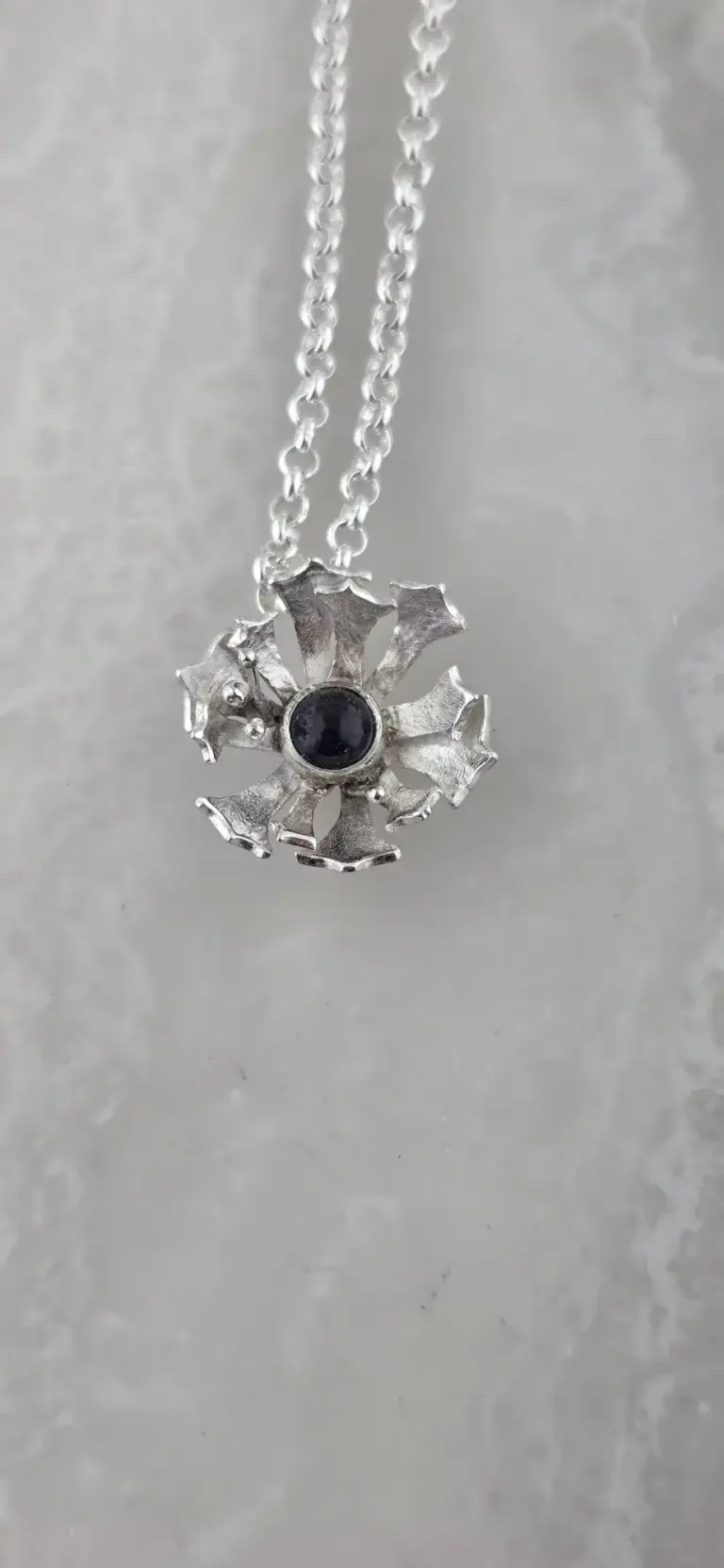 Silver necklace with a flower-shaped pendant featuring a dark gemstone at the center, hanging on a chain.