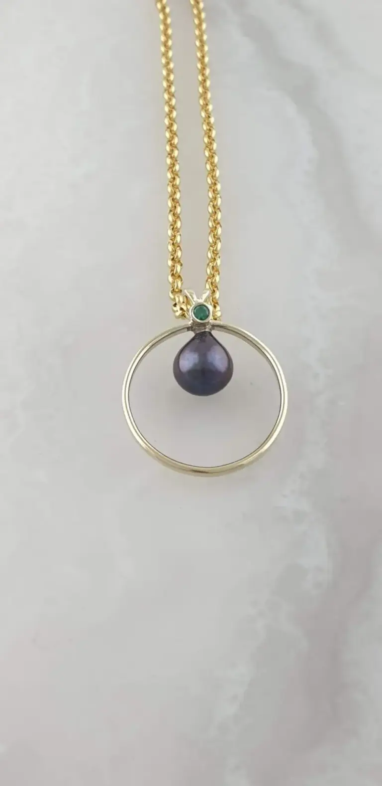Gold necklace with a circular pendant featuring a dark purple pearl and a small green gemstone set above the pearl.