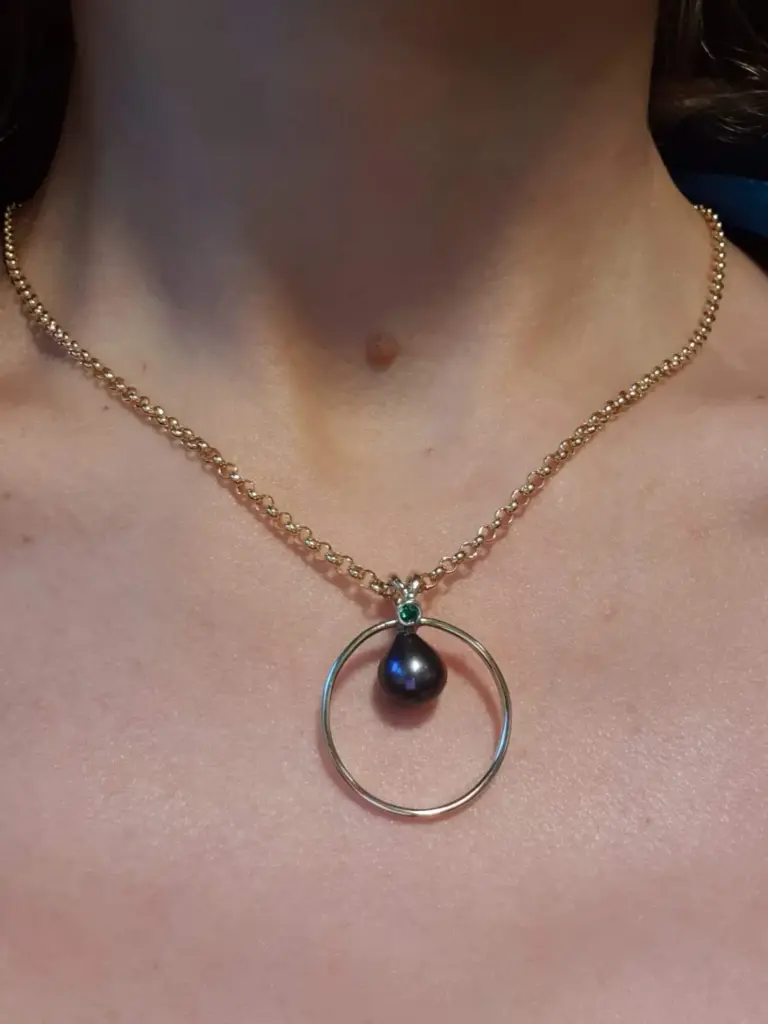 A close-up of a woman's neck and collarbone, showing a gold chain necklace with a large black pearl pendant and a small green gemstone detail, against a pink background.