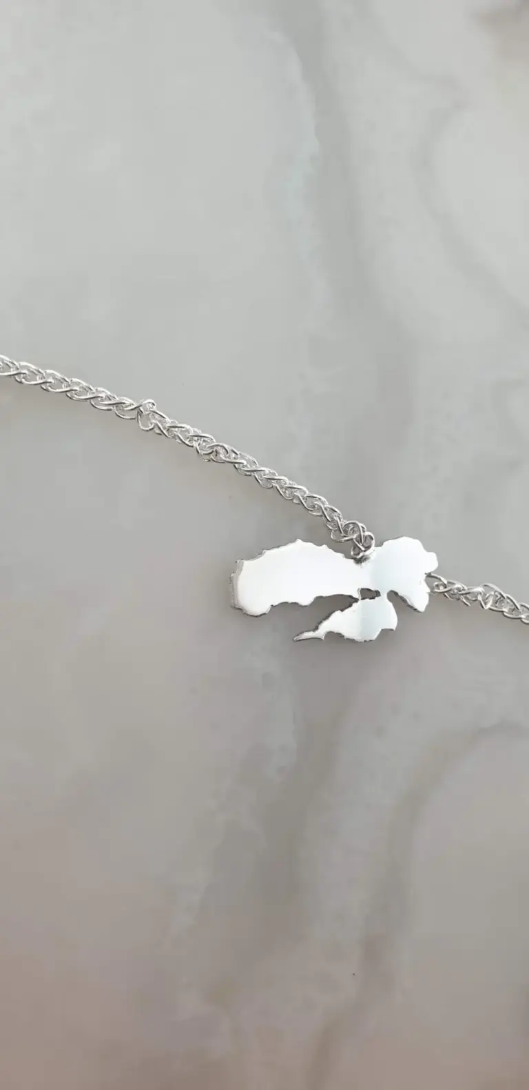 A silver necklace with a pendant in the shape of the state of Alaska, resting on a light-colored surface.