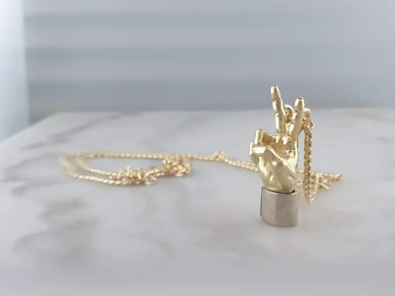 Gold jewelry including a hand-shaped pendant and a chain, displayed on a white stand against a light background.