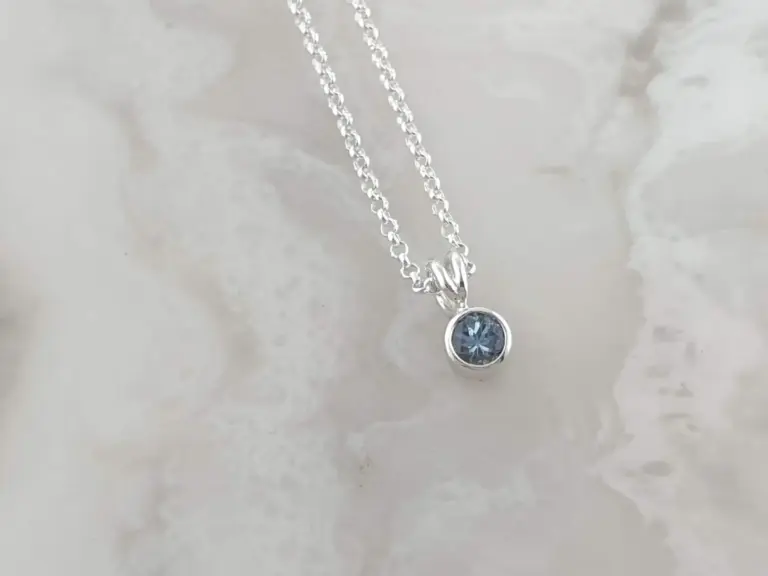 A silver necklace with a round blue gemstone pendant hangs against a light-colored surface.