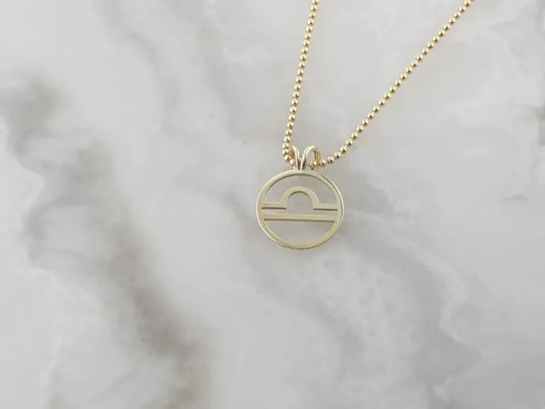 Gold necklace with a circular pendant featuring a stylized design, hanging on a beaded chain against a white, textured background.