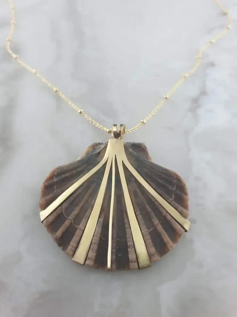 A decorative necklace with a gold chain and a pendant resembling a fan or a shell, resting on a surface with a shell underneath.