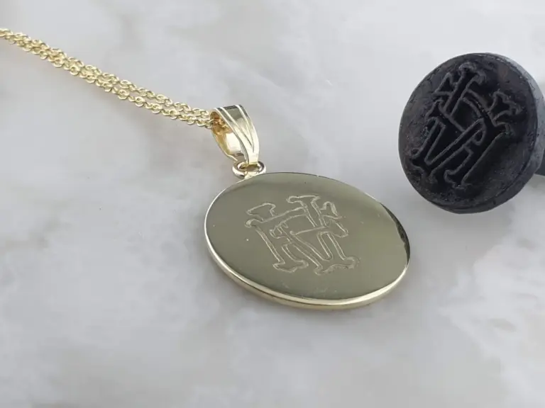 Close-up of a gold necklace with a engraved pendant and a black stone stamp with embossed text on a light surface.