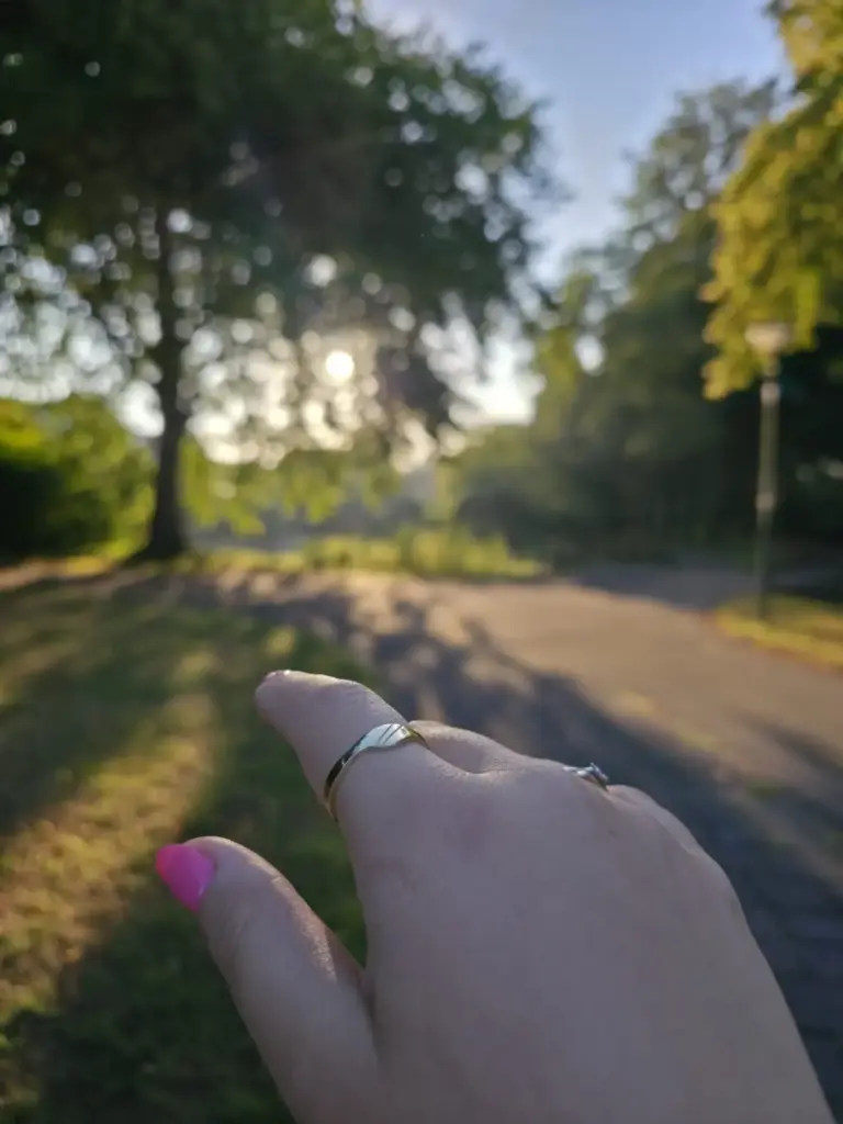 A person with pink nail polish on their thumb and multiple rings on their fingers, reaching out towards the sunlight through a park with large trees in the background.