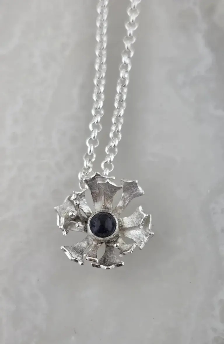 Silver necklace with pendant shaped like a flower with jagged edges and a central black stone, hanging on a silver chain against a light, neutral background.