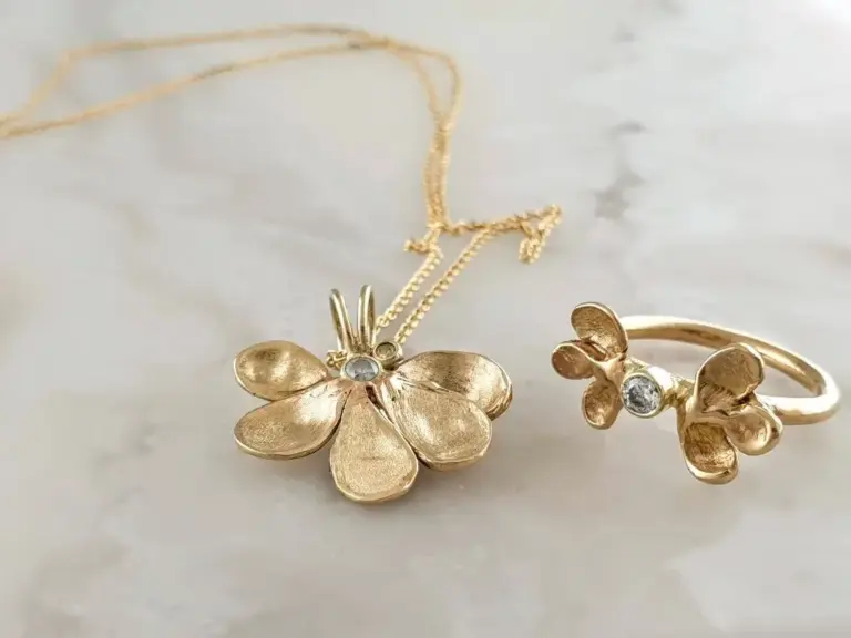 Gold flower-shaped necklace and ring with small clear gemstones, on a white marble surface.