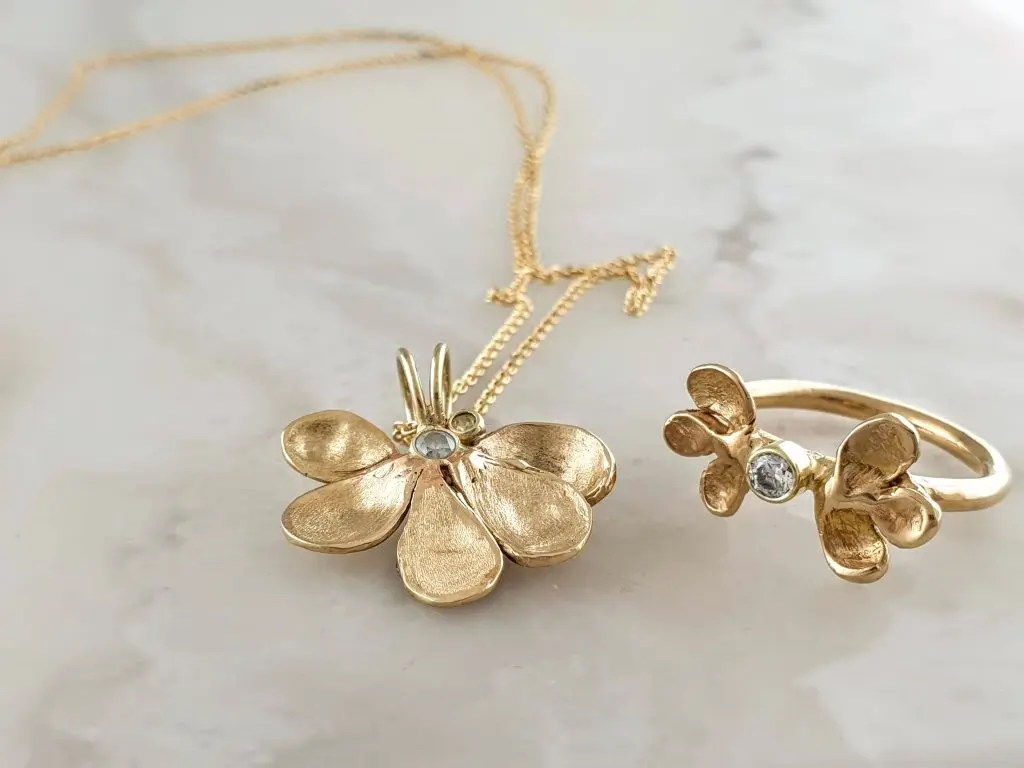 Gold flower-shaped necklace and ring with small clear gemstones, on a white marble surface.