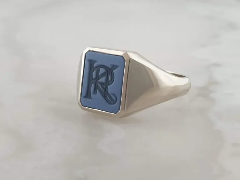 Close-up of a silver ring with a blue rectangular face engraved with the letter 'R' in cursive, resting on a plain white surface.
