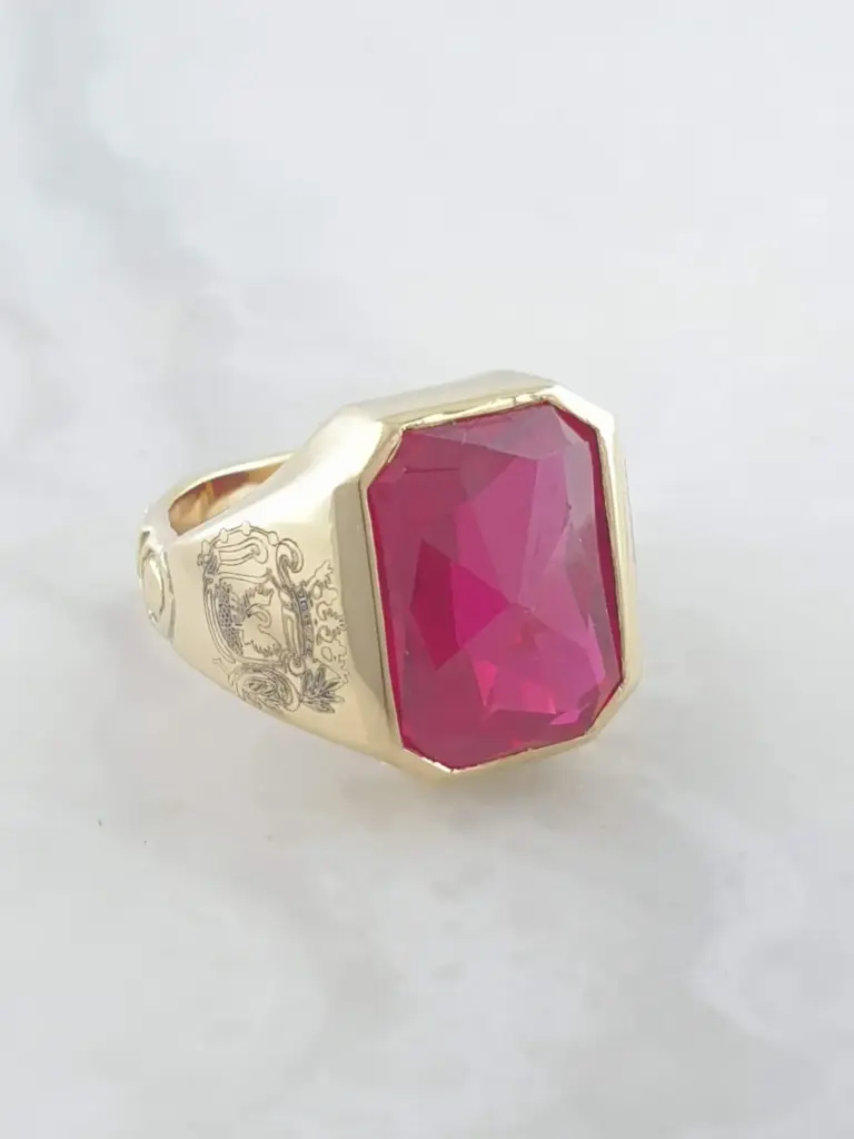 A gold ring with an elaborate engraved design on the band, featuring a large, rectangular, faceted pink gemstone.