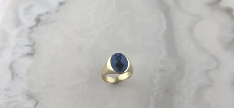 Close-up of a gold ring with an oval blue stone featuring a black bear engraving, placed on a white marble surface.