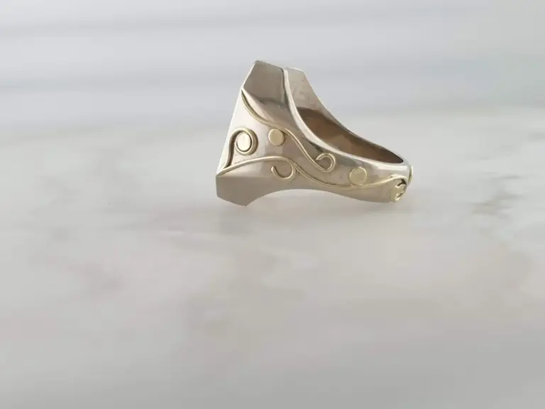 A silver ring with decorative gold swirls and circular designs, positioned on a plain, gray surface.