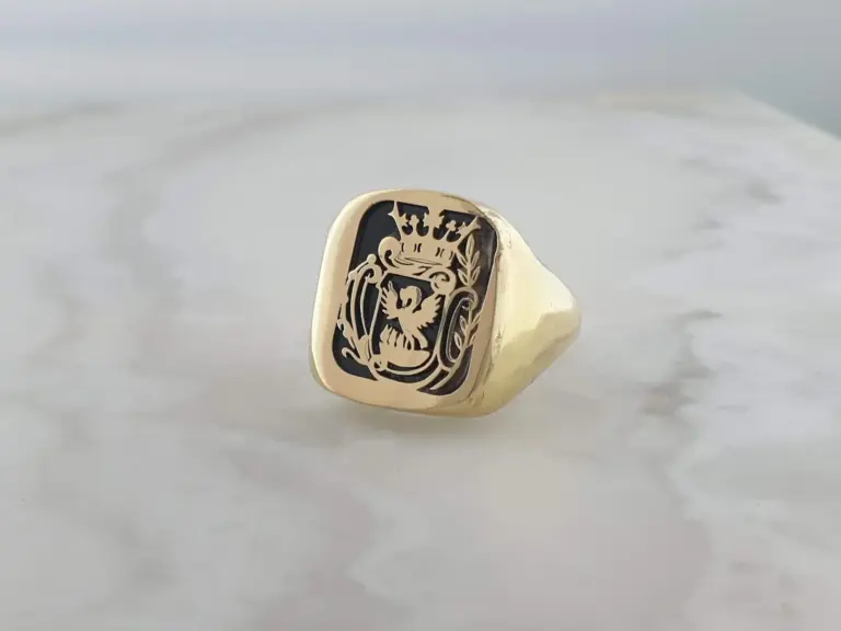 Close-up of a gold ring with an engraved crest or emblem, featuring a shield, a crown, and ornate decorations.