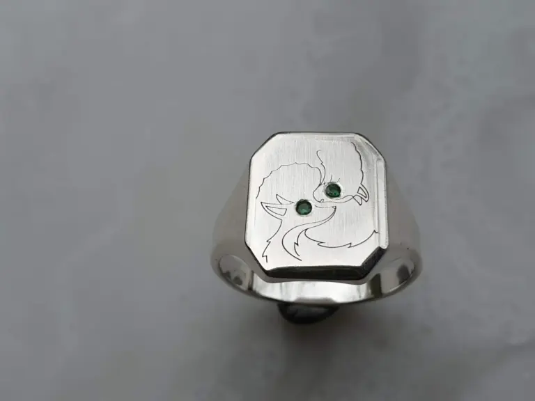 A silver ring featuring a design of cartoon cat faces with green gemstone eyes engraved on the top.