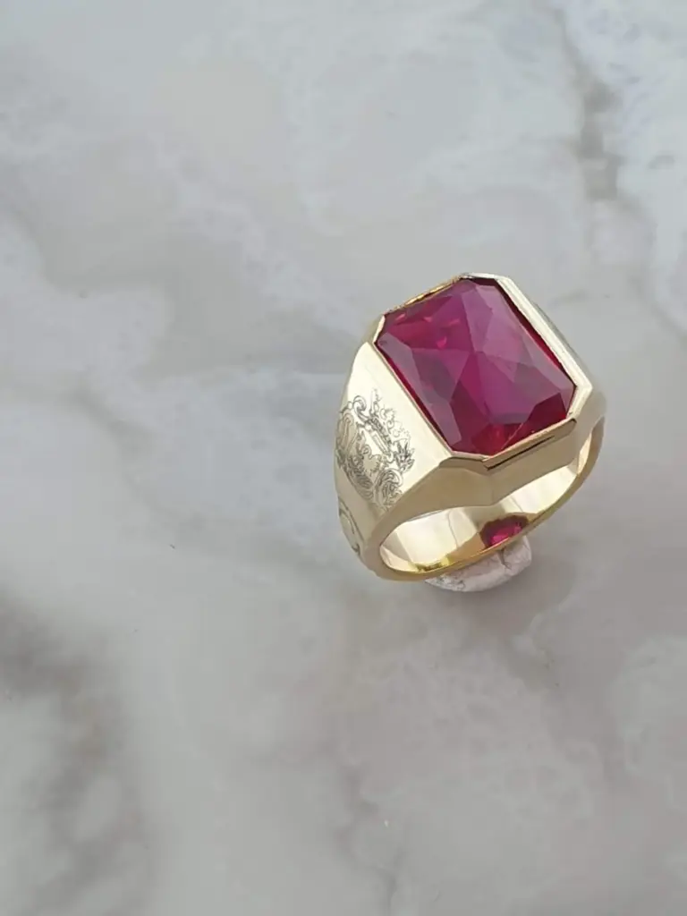 A gold ring with a large rectangular red gemstone and intricate engraved details on the sides, placed on a white textured surface.