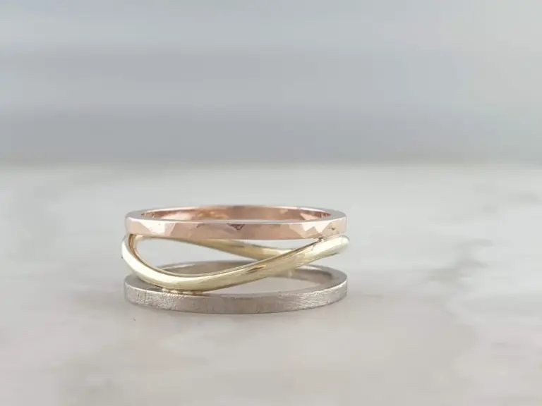 Stack of four rings with various textures and metallic finishes, including rose gold, gold, silver, and textured silver, arranged on a white surface with a plain gray background.