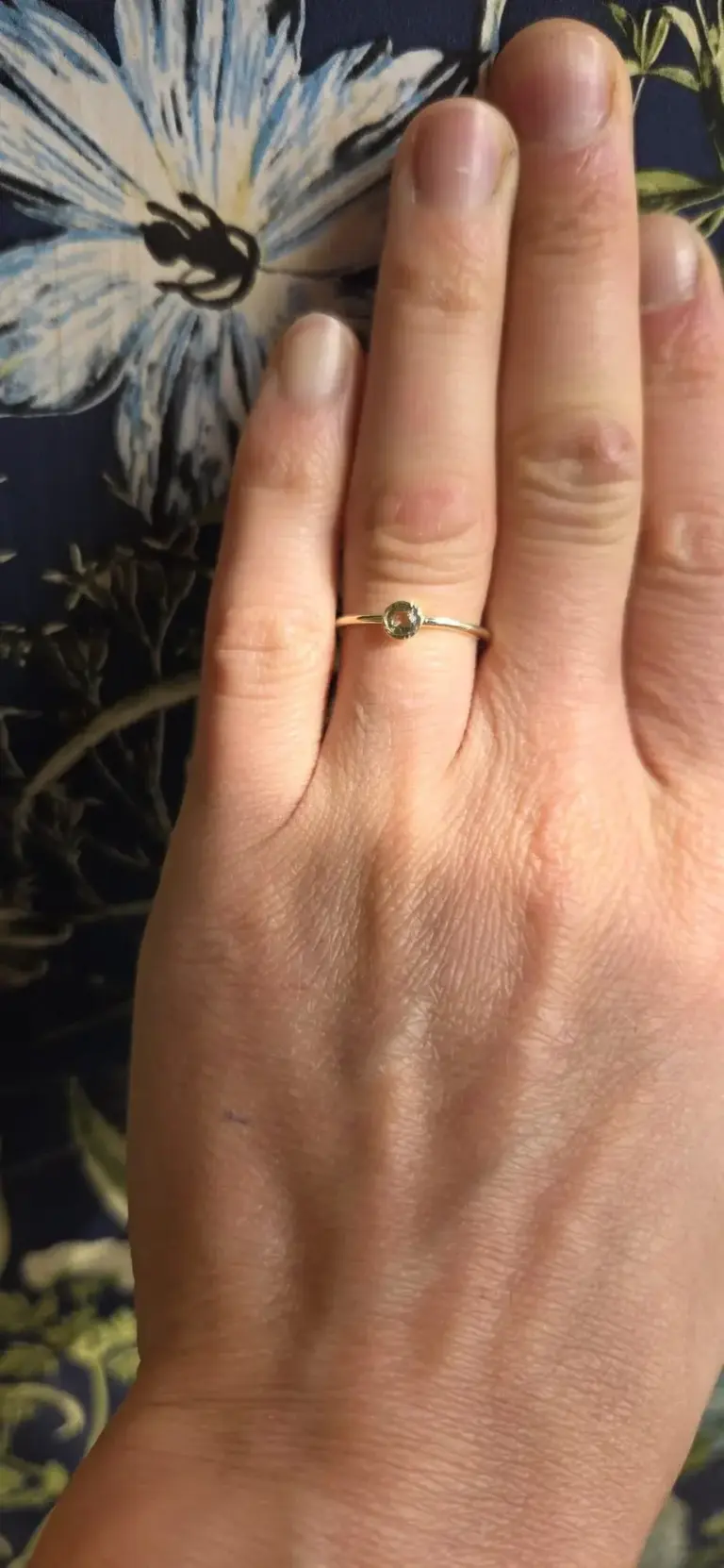 A person's hand with a ring featuring a round, clear gemstone on the ring finger, against a background with floral patterns.