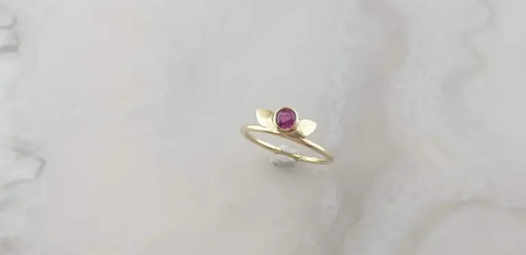 Gold ring with a pink gemstone, displayed on a small white stand against a light textured background.
