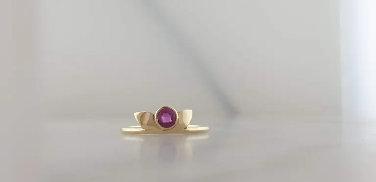 A gold ring with a large pink gemstone in the center, resting on a flat surface against a light, blurred background.
