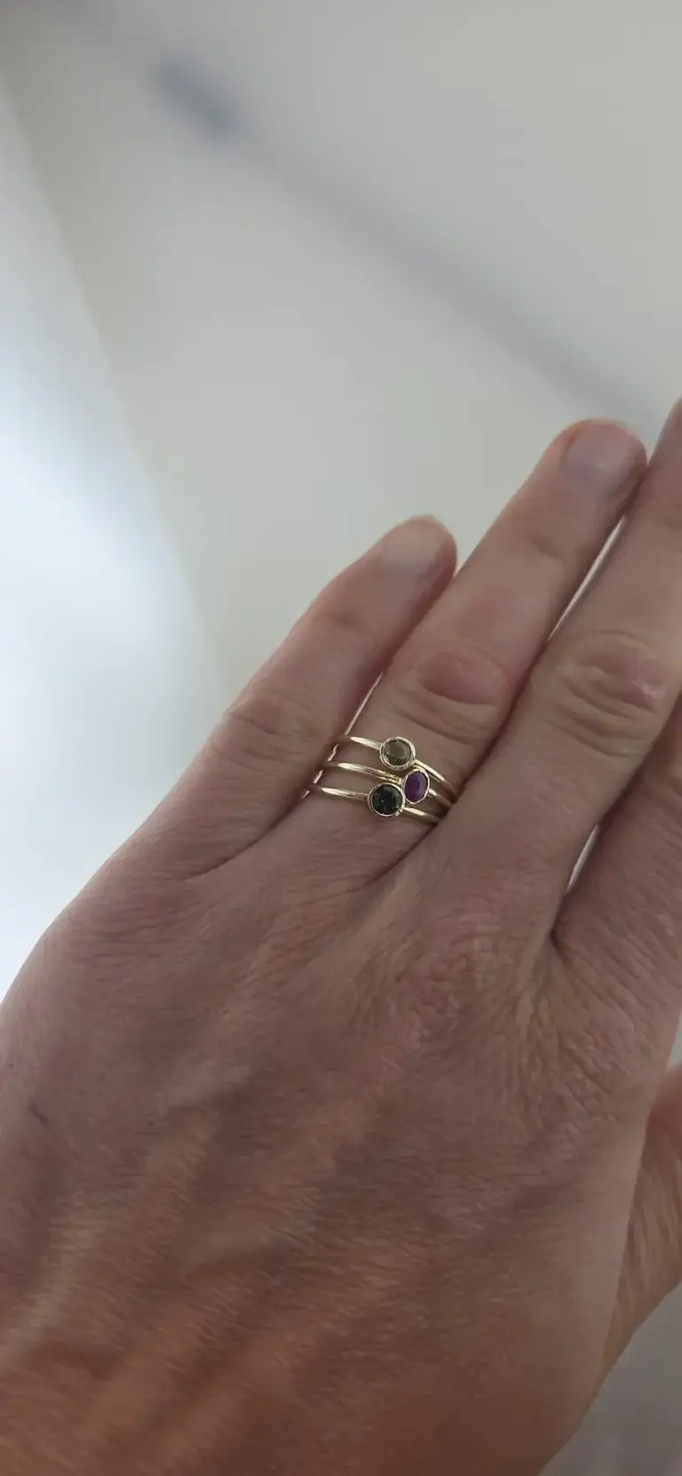 Close-up of a hand wearing a gold ring with three small gemstone settings featuring black, purple, and clear stones.