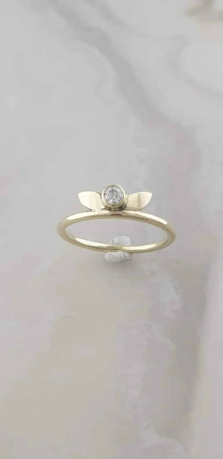 A gold ring with a small round diamond in a bezel setting, flanked by two gold leaves, displayed on a white surface with a blurred background.