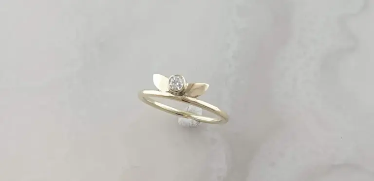 Gold ring with leaf design and a centered diamond, displayed on a small white stand against a light background.