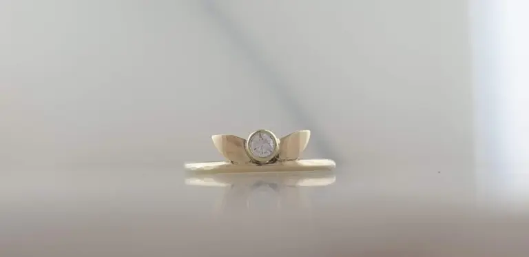 Close-up of a gold ring with a prominent diamond in the center, resting on a reflective surface with a blurred light-colored background.