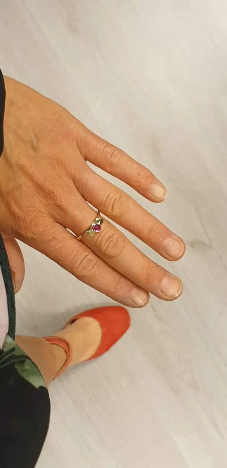 A person's hand wearing a gold ring with a pink gemstone, with visible wrinkles, red-painted nails, and a facial mask on their face. The background is a light-colored wall and part of an orange shoe is visible.