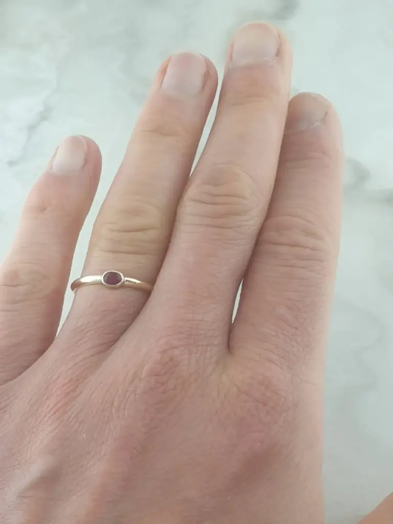 Close-up of a person's left hand displaying their fingers and a ring with a red stone on the ring finger, against a soft, light background.