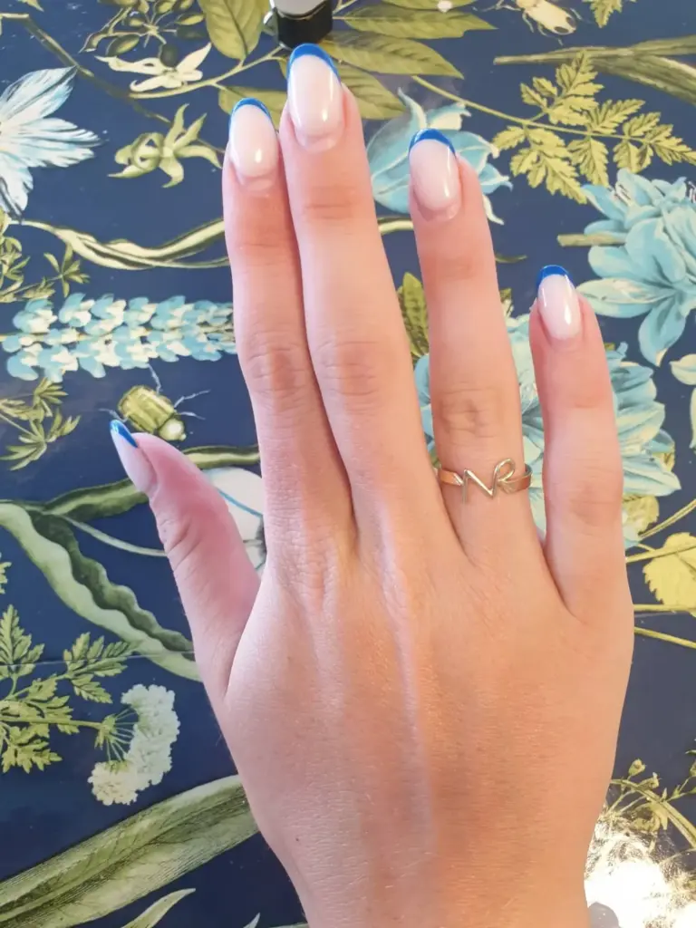 Close-up of a hand with a gold ring reading 'N', showing manicured nails with clear polish and blue French tips, against a floral wallpaper background.