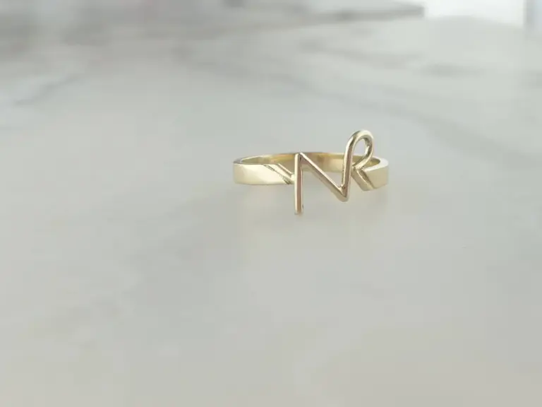 A gold ring with a letter 'N' shaped design resting on a reflective surface.