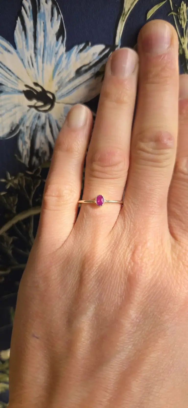 A hand wearing a gold ring with a pink gemstone on the ring finger, positioned against a dark background with a floral pattern.