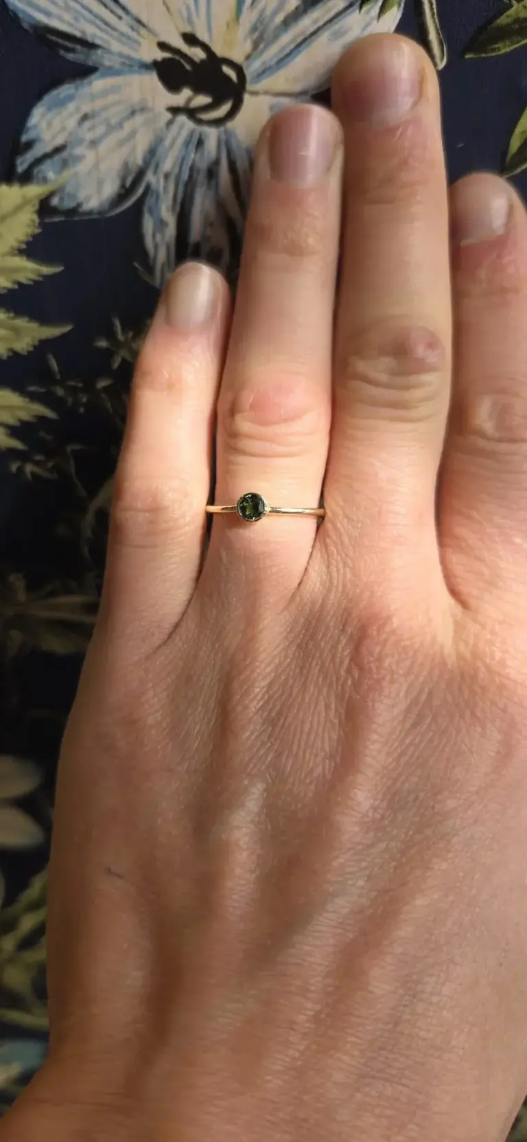 Close-up of a hand with a gold ring featuring a small round green gemstone, resting on a patterned fabric background.