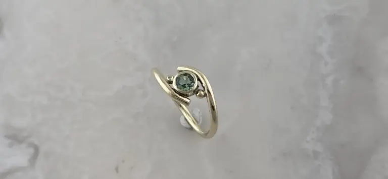 A gold ring with a green gemstone, set in a modern, swirl design, on a light gray surface.