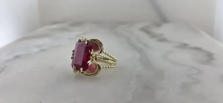 Gold ring with a large, rectangular pink gemstone set in a decorative setting, displayed on a white textured surface.