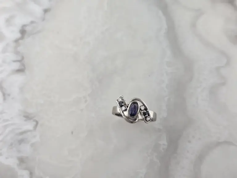 Close-up of a silver ring with a large purple gemstone and smaller clear stones, resting on a textured surface.