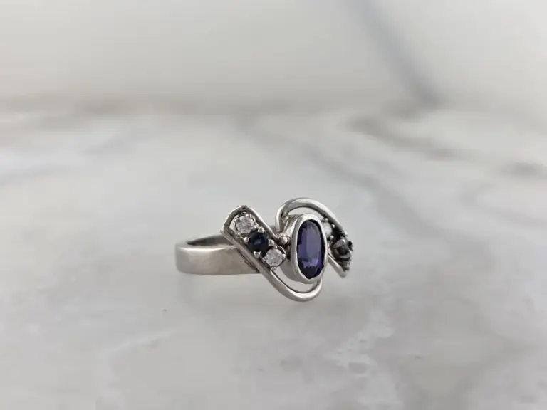 A silver ring featuring a large purple gemstone in the center, accompanied by smaller clear and dark gemstones, resting on a light-colored surface.