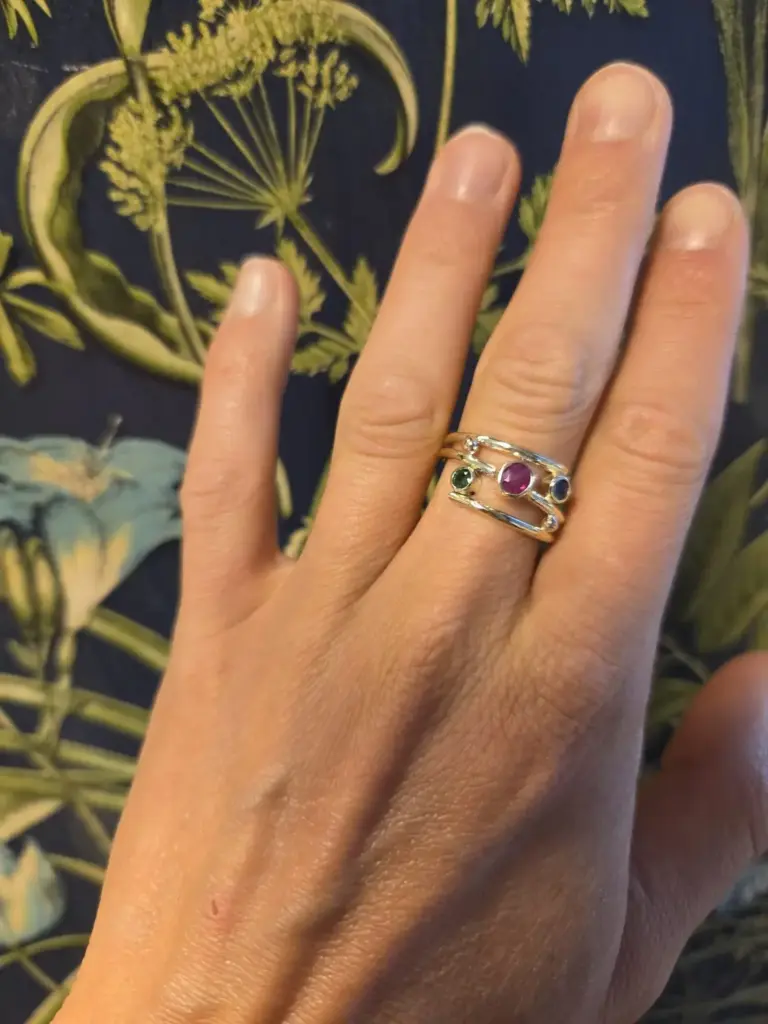 A person's hand wearing three silver rings with colorful gemstones, resting on a dark fabric with yellow-green floral patterns.