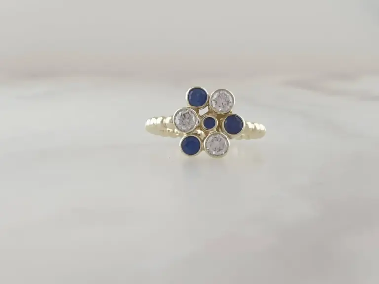 A gold ring featuring alternating white and blue gemstones, arranged in a cluster, on a reflective surface.